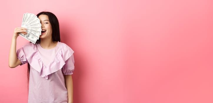 Happy rich asian woman showing money and smiling, shopping with cash, standing in dress against pink background.