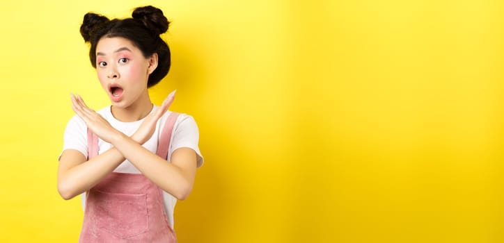 She says no. Asian beauty girl with makeup, showing cross sign, stop and reject offer, standing disappointed on yellow background.