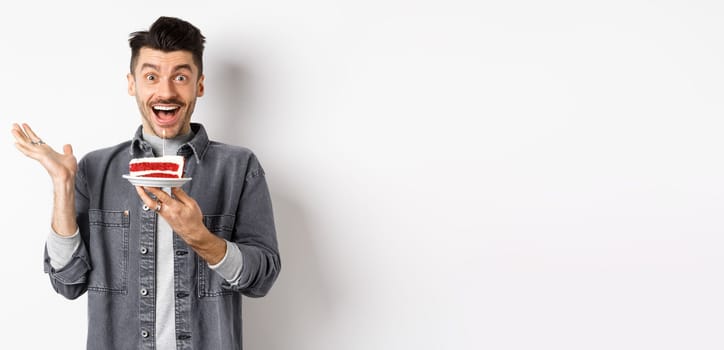 Excited man celebrating birthday, looking happy, holding cake with candle, making wish, standing happy against white background.