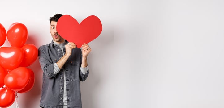 Cute young man hiding face behind big Valentine heart card, secret admirer looking at camera with amazement, saying wow, standing near romantic balloons on white background.
