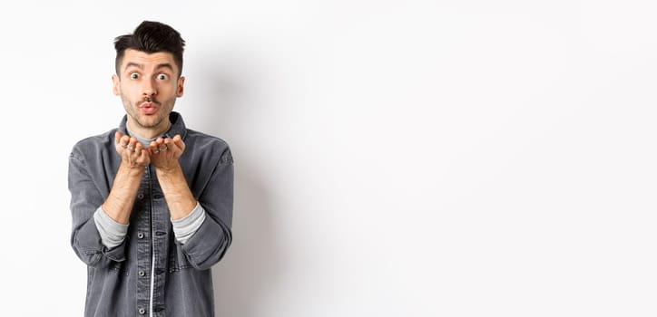 Romantic handsome man blowing air kiss, looking silly at camera, standing in trendy clothes on white background.