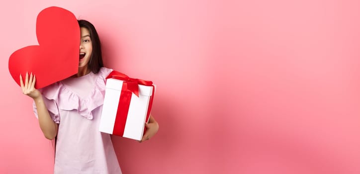Valentines day concept. Happy young girl asian holding romantic gifts, red heart card from lover and present box, looking amazed at camera, standing in dress on pink background.