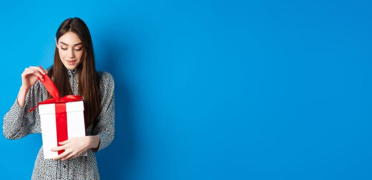 Valentines day. Cute young woman open box with gift, take-off ribbon from present and smiling intrigued, standing on blue background.