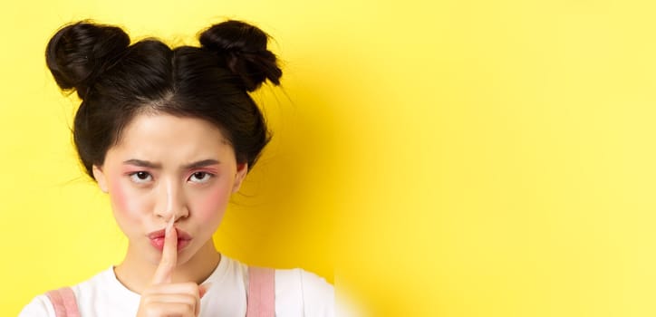 Close up portrait of angry asian girl shushing, press fingers on lips and tell be quiet, scolding loud person, need silence, telling big secret, standing on yellow background.