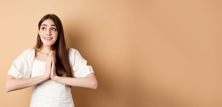 Hopeful young girl begging god and looking dreamy at upper left corner, praying or making wish, standing on beige background.