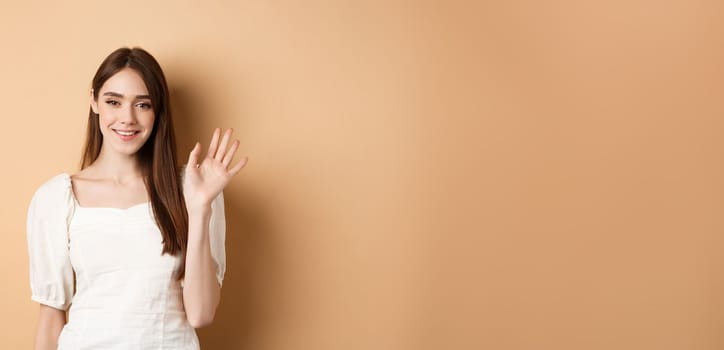 Friendly smiling woman say hello, waiving hand to greet you, standing cheerful on beige background.