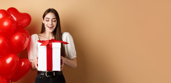 Happy woman open Valentines day gift, smiling excited and looking at present box, standing near red heart balloons on beige background.