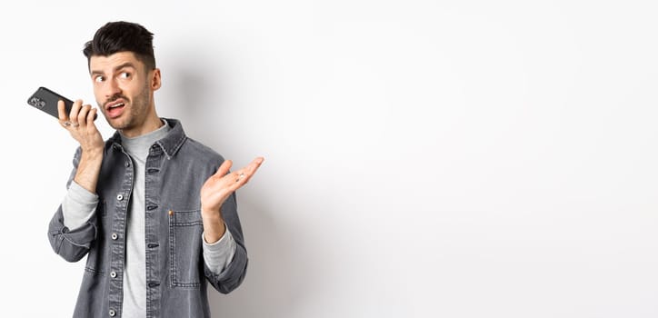 Stylish handsome man talking on speakerphone, record voice message with smartphone near lips, discuss something and looking aside at empty space, standing on white background.