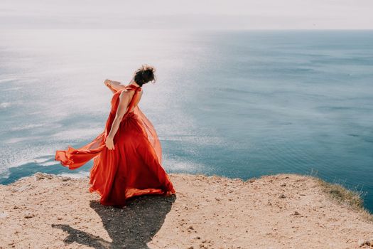 Side view a Young beautiful sensual woman in a red long dress posing on a rock high above the sea during sunrise. Girl on the nature on blue sky background. Fashion photo.