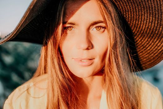 Portrait of happy young woman wearing summer black hat with large brim at beach on sunset. Closeup face of attractive girl with black straw hat. Happy young woman smiling and looking at camera at sea