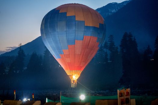 Hot air balloon with fire heating air in wicker basket with himalaya mountains in background showing this adventure in kullu manali valley India