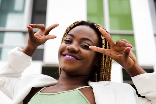 Head shot of Black woman with braided hair looking at camera showing peace sign with hands. Lifestyle concept.