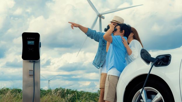 Concept of progressive happy family enjoying their time at wind farm with electric vehicle. Electric vehicle driven by clean renewable energy from wind turbine generator for charging station.