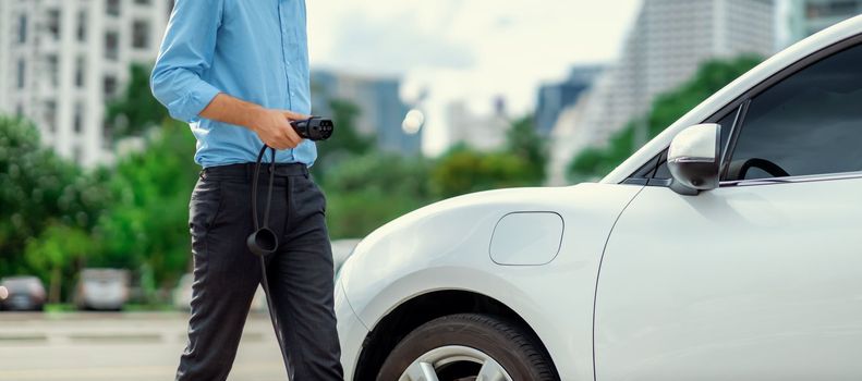 Closeup progressive man holding EV charger plug from public charging station for electric vehicle with background of residential building as concept eco-friendly sustainability energy car concept.