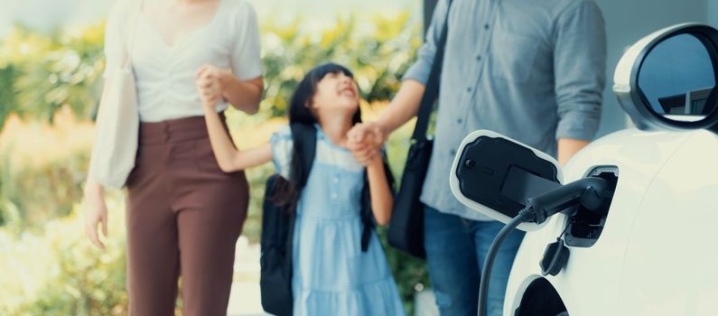 Focus closeup electric vehicle recharging battery from home electric charging station with blurred family in background. Renewable clean energy car for progressive eco awareness lifestyle concept.