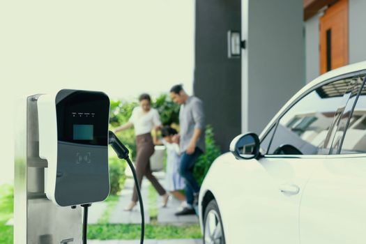 Focus closeup electric vehicle recharging battery from home electric charging station with blurred family in background. Renewable clean energy car for progressive eco awareness lifestyle concept.