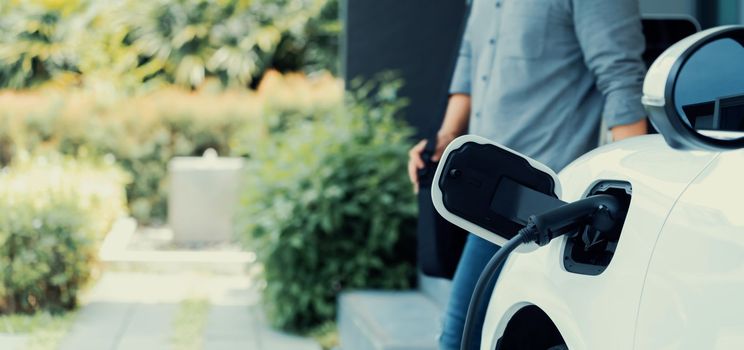 Focus electric car charging at home charging station with blurred progressive man walking in the background. Electric car using renewable clean for eco-friendly concept.