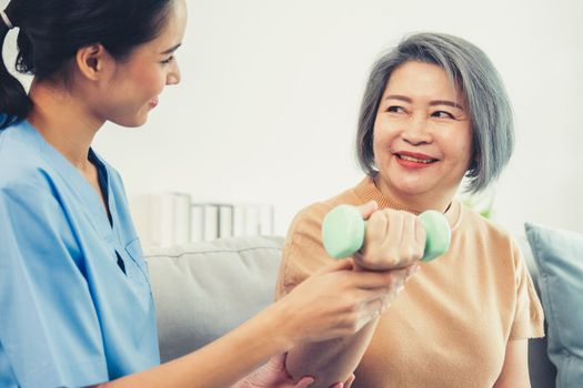 Caregiver helping contented senior woman exercise with dumbbell at home. Rehab for elder, senior medical care.