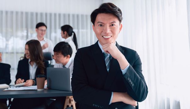 Portrait of focus young successful confident male manager, executive wearing business wear in harmony office arm crossed with blurred meeting background of colleagues, office worker.
