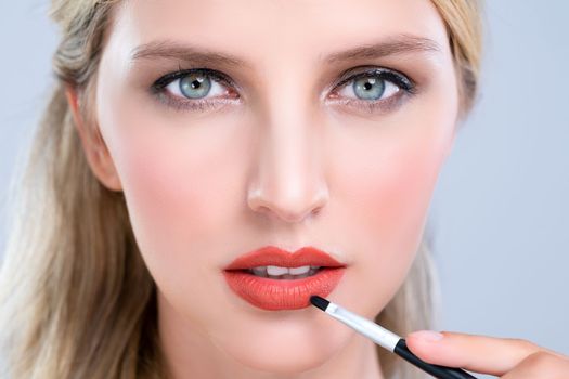 Closeup beautiful young woman with flawless healthy skin and natural makeup putting alluring fashion glossy red lipstick on her lip with lip brush in isolated background.