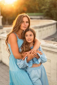 Portrait of mother and daughter in blue dresses with flowing long hair against the backdrop of sunset. The woman hugs and presses the girl to her. They are looking at the camera