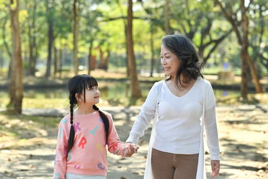 Joyful little girl and grandmother walking in park surrounded by green trees at sunlight morning. Family, generation concept.