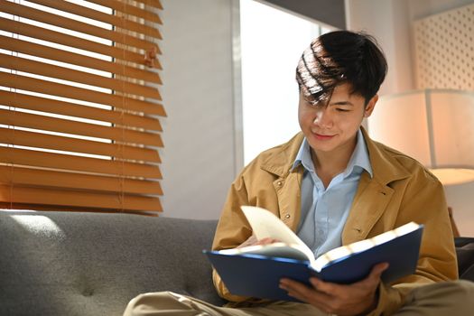 Relaxed millennial man reading book on comfortable couch, enjoying leisure weekend at home.