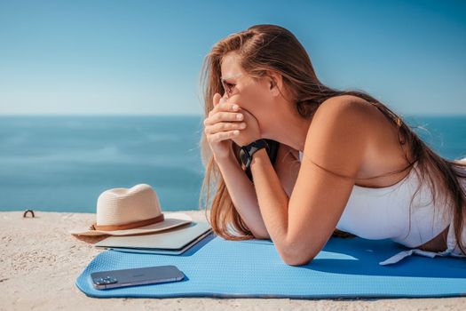 Successful business woman in yellow hat working on laptop by the sea. Pretty lady typing on computer at summer day outdoors. Freelance, travel and holidays concept.