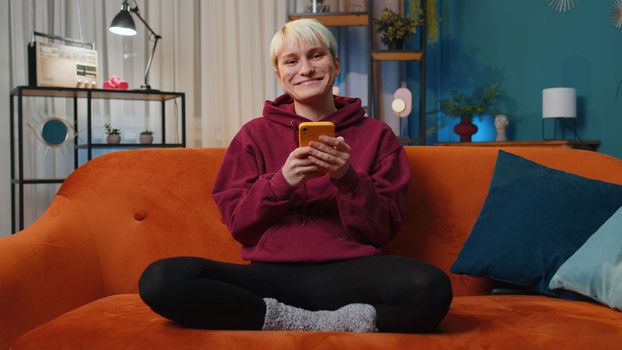 Woman with short blonde hair sitting on sofa uses mobile phone smiles at night home apartment. Girl texting share messages content on smartphone social media applications online, watching relax movie