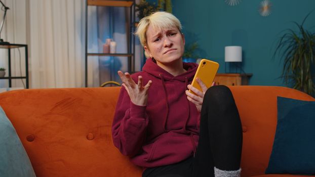 Sad woman with short blonde hair use smartphone typing browsing, loses becoming surprised sudden lottery results, bad news, fortune loss, fail, deadline. Young adult girl at evening home room on couch