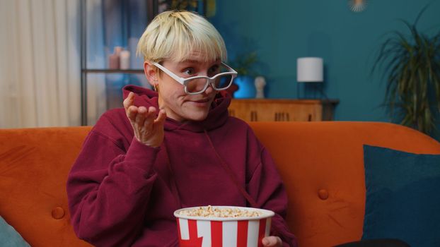 Excited young woman sits on sofa eating popcorn snacks and watching interesting TV serial, sport game, film, online social media movie content at home. Girl enjoying evening entertainment. Lifestyles