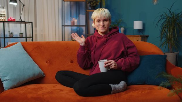 Young woman with short blonde hair smiling friendly at camera and waving hands gesturing hello, hi, greeting or goodbye, welcoming with hospitable expression. Girl indoors at home in room on couch