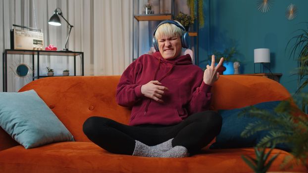 Happy rocker woman with short hair in wireless headphones relaxing at home dancing on couch listening energetic disco dancing music, playing on imaginary guitar. People weekend leisure activities