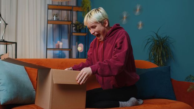 Happy surprised winner woman unpacking delivery parcel at home. Smiling satisfied girl shopper online shop customer opening cardboard box receiving purchase gift headphones by fast postal shipping