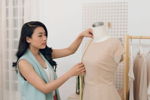 Young Asian female fashion designer taking measurements on mannequin in her studio