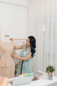 Young Asian female fashion designer taking measurements on mannequin in her studio