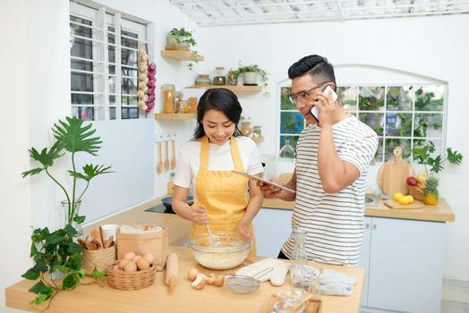 Young asian man and woman together cooking cake and bread with egg, looking menu from tablet in the flour happy relaxing in at home