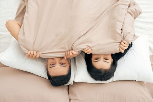Funny married couple lying in bed and hiding under white blanket, looking at camera with eyes full of joy.