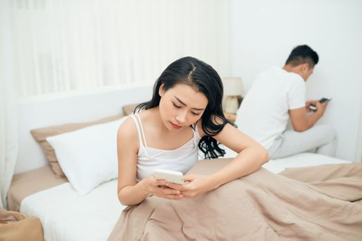 sad view of young married couple using their mobile phone in bed ignoring each other as strangers in relationship and communication problems