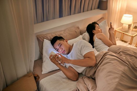 Young couple in bed using phone lying backs to each other.