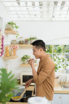 Attractive young man is cooking on kitchen with laptop on table while talking on smart phone.