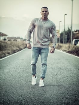 One handsome young man in urban setting walking along street, wearing sweater and jeans