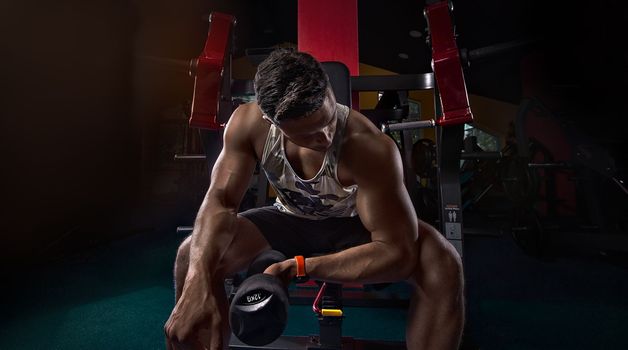 athlete strains biceps in the gym with dumbbells, creative photo, contours, muscles stand out with a watch on his arm, in an empty gym, beautiful body, physique, relief, sexy guy