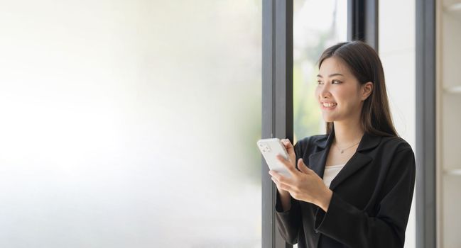 Portrait of elegant successful asian business woman speaking by smartphone and smiling happily while standing against window in office