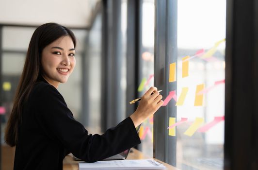 Young smiley attractive, businesswoman using laptop and post it notes in glass wall to writing strategy business plan to development grow to success..