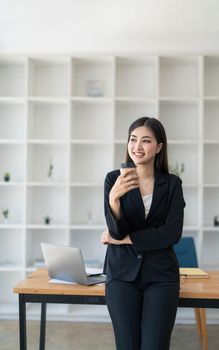 Portrait of young confident woman holding coffee cup, standing at office and looking outside. Successful businesswoman standing in office with copy space...