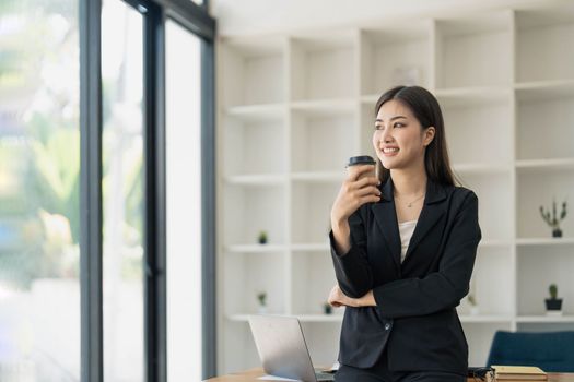 Portrait of young confident woman holding coffee cup, standing at office and looking outside. Successful businesswoman standing in office with copy space...