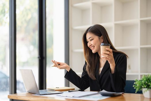 Beautiful young Asian businesswoman smiling holding a coffee mug and laptop working at the office...