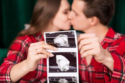 Close-up on the hands of a man and a woman holding an usg photo of a baby. In the background are the blurred faces of the kissing couple and a dark green curtain.