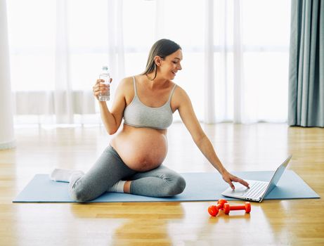 Portrait of a young happy pregnant woman doing exercise and looking at a laptop and drinking water at home or in a gym